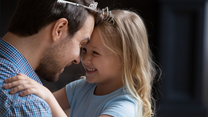 Overjoyed little preschooler girl child touch foreheads look in eyes playing with loving young dad wearing toy crown, happy father and small daughter have fun playing together funny games at home