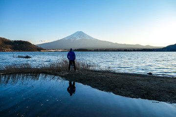 Fototapeta na wymiar A man walking at the side of Kawaguchiko Lake and watching Mt Fuji, Japan. Reflection of the man in the water. Top of volcano covered with snow. Exploring new places. Soft sunset colors. Calmness