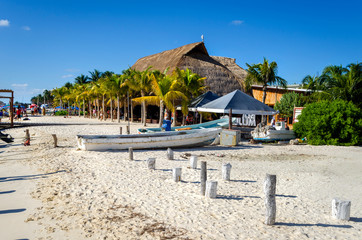 Fototapeta premium A Caribbean beach with a fishing boats tied up outside a coffee shop showing beep blue sky and a line of palm trees running along the shore side. .