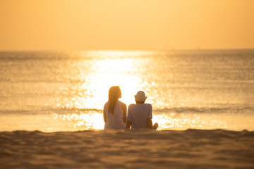 couple sit on sand looking sunset in sea together.