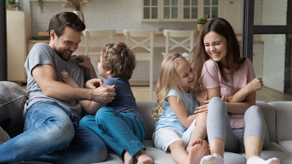 Playful young parents have fun play with happy little kids tickle laugh sitting on couch in living room, overjoyed family enjoy funny childish activity or game with small children on weekend at home