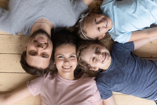Top view close up portrait of happy young family with little children lying on warm wooden floor posing for picture together, smiling parents and small kids relaxing at home look at camera