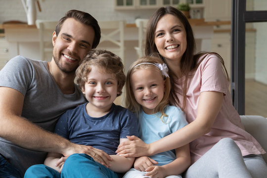 Close up headshot portrait of smiling young parents hug cuddle posing with little preschooler kids at home, happy family with children look at camera embracing relax enjoying weekend together