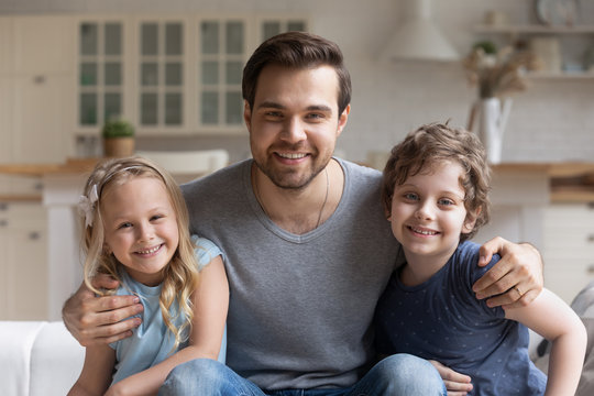 Close up portrait of smiling young father and small kids sit on couch look ta camera posing, happy dad parent hug excited cute little preschooler boy and girl children relaxing at home together