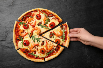 Woman taking piece of delicious seafood pizza at black table, top view