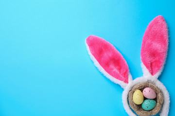 Headband with Easter bunny ears and dyed eggs in nest on light blue background, flat lay. Space for text