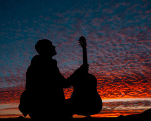 Silhouette of male musician sitting holding guitar on scenic violet to red sunset landscape