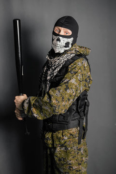 Unknown man, a robber in a balaclava with a skull, in camouflage uniform and body armor with a baseball bat in his hands. Studio photo on a gray background.