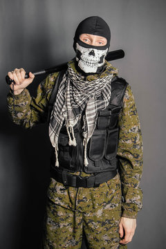 Unknown man, a robber in a balaclava with a skull, in camouflage uniform and body armor with a baseball bat in his hands. Studio photo on a gray background.