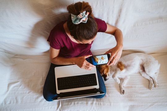 young woman working on laptop at home, sitting on the couch, taking a picture with mobile phone of her cute small dog besides. Technology and pets concept