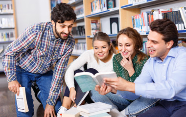 Group of happy students studying together in library, sitting on floor on background with bookshelves