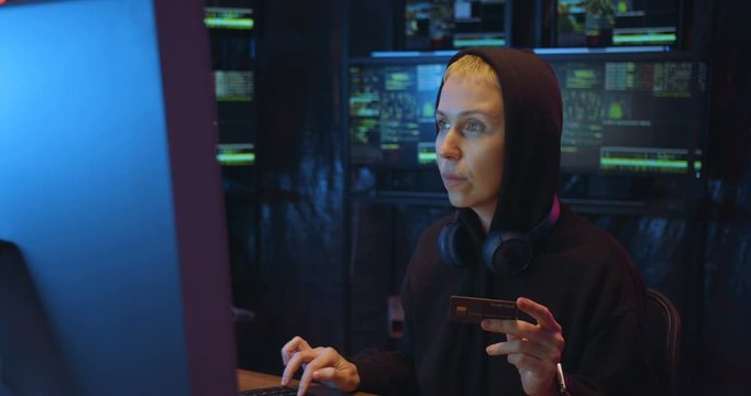 Caucasian woman in hood typing credit card number on keyboard, shopping online at big computer screen in dark IT monitoring room. Female hacker buying and paying via internet. Financial cyber crime.