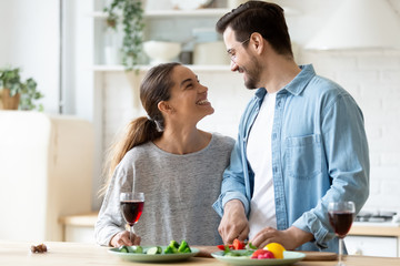 Obraz na płótnie Canvas Happy young couple cooking dinner in kitchen at home together, smiling husband cutting fresh vegetables, preparing salad, beautiful wife holding wine glass, enjoying romantic moment, having fun