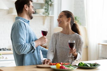 Young loving couple having fun in kitchen, drinking red wine, holding glasses, laughing beautiful wife and husband chatting, preparing salad, cooking dinner, enjoying free time together