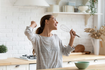 Excited funny girl singing into whisk, having fun in modern kitchen at home, happy girl holding...