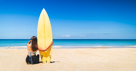 Women Caucasian hold a yellow surfboard on the beach. Women are viewing waves to surf the waves in good weather and clear skies during the summer in holiday and activity concept with copy space.