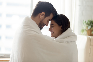 Happy loving couple wrapped in warm blanket touching foreheads with closed eyes and hugging, standing in kitchen at home, smiling beautiful wife and handsome husband enjoying tender moment