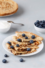 Four thin pancakes on white plate with blueberries and sour cream. Grey background.