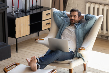 Calm young man relaxing with laptop on cozy armchair, peaceful male with closed eyes and hands...