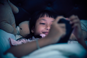 Asian child girl is playing smartphone on the bed at night.