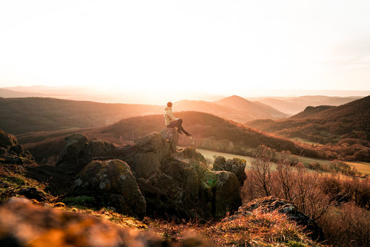 Adult tourist in black trousers, jacket sit on cliff's edge and looking to misty hilly valley bellow. Hker relax and thinking alone in nature.