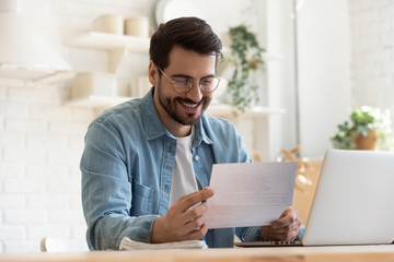 Smiling young man reading good news in paper notification, sitting at table with laptop, happy satisfied male wearing glasses holding document, job promotion, loan approval, great exam result - obrazy, fototapety, plakaty