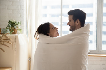 Happy loving couple wrapped in white warm blanket hugging, standing in kitchen at home, smiling...