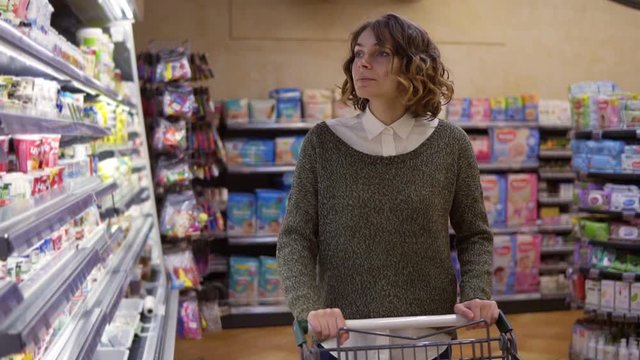 Food, Health Concept - Woman in a supermarket standing in front of the freezer shelfs and choose buying a bottle of jogurt, taking one and put it to the cart . Slow motion