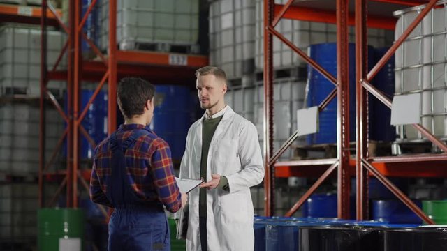 Tilt down medium shot of middle aged engineer in white coat explaining something to young warehouse worker with clipboard in factory storehouse