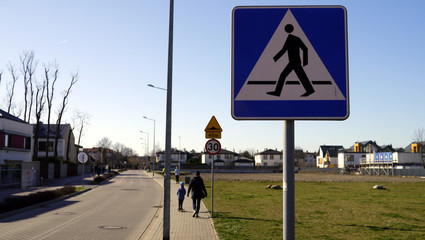 Znak przejście dla pieszych. W tle idzie matka z dzieckiem i widać znak ograniczenia prędkości...