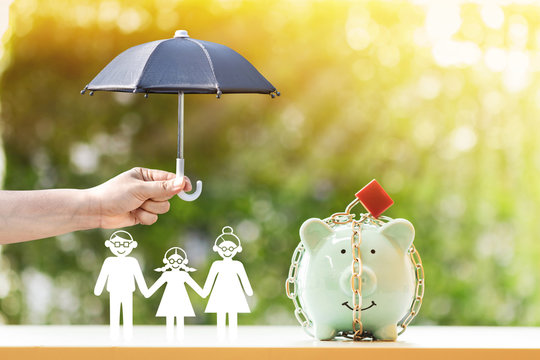 Piggy bank with security and lock and family and woman hand hold the black umbrella for protect on sunlight in the public park, to prevent for asset and saving money for buy health insurance concept.