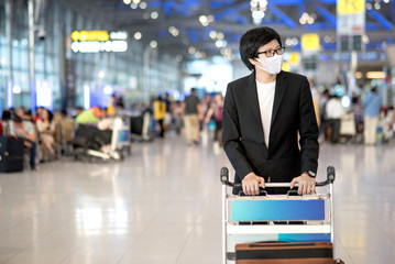 Asian businessman wearing suit and face mask walking with airport trolley and suitcase luggage in airport terminal. Coronavirus (COVID-19) outbreak prevention. Health awareness for pandemic protection
