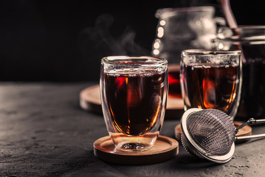 Cup of freshly brewed black tea, dark moody, tea ceremony. Hot water is poured from the kettle into a flass cup on wooden tray, black concrete background.