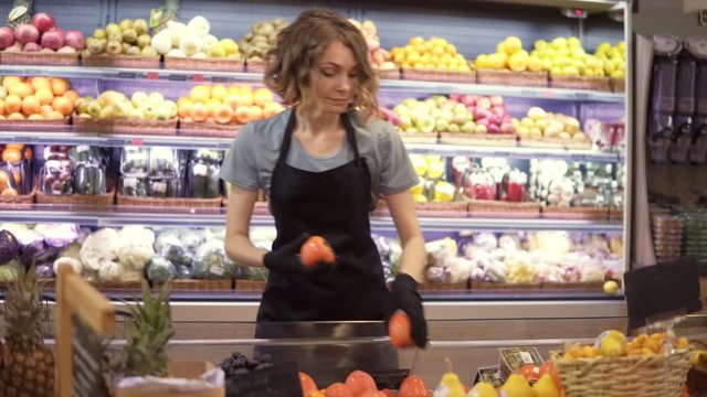 Portrait of female shop assistant in gloves and black apron filling up fruits and vegetables storage stands with assorted organic groceries in supermarket. Slow motion