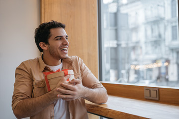 Portrait of young emotional asian guy with birthday present in hand. Happy handsome Indian man holding gift box, sitting in modern cafe, looking at window and smiling