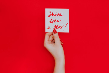 Note in a female hand with manicure, red nails. "shine like a star!" sign. Background red