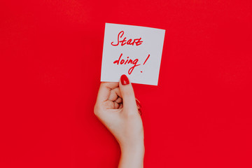 Note in a female hand with manicure, red nails. "Start doing!" sign. Background red