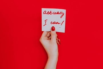 Note in a female hand with manicure, red nails. "Actually i can!" sign. Background red