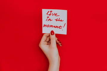 Note in a female hand with manicure, red nails. "Live in the moment!" sign. Background red