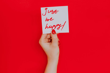 Note in a female hand with manicure, red nails. "Just be happy!" sign. Background red