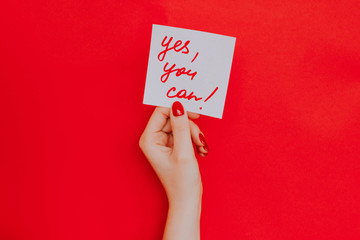 Note in a female hand with manicure, red nails. "Yes you can!" sign. Background red
