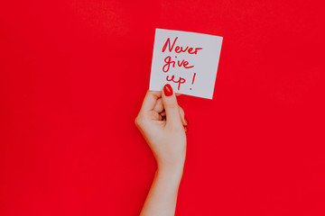 Note in a female hand with manicure, red nails. "Never give up!" sign. Background red