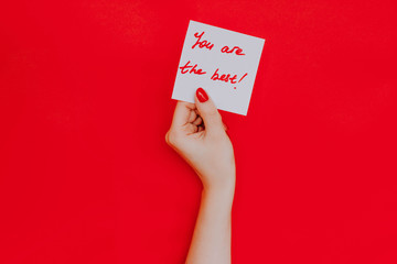 Note in a female hand with manicure, red nails. "You are the best!" sign. Background red