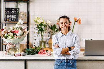 Confident female florist standing with her arms crossed and looking at camera. Happy flower shop owner standing at counter. - obrazy, fototapety, plakaty
