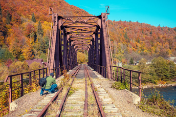 Old railway on the bridge over the mountain river in autumn. A man takes a picture of the railway