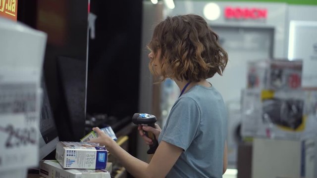 Curly haired concentrated woman seller-consultant in household department carry and scan by barcode stationery goods in the boxes, standing in front the screen. Slow motion. Side view