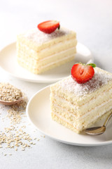 Dessert. White biscuit cake with cream and coconut shavings on white plate with spoon. Strawberry on a light background. Background image, copy space