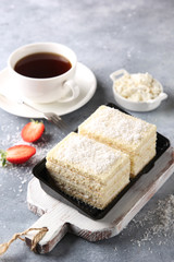 Dessert. Biscuit cake with cottage cheese, cream and coconut shavings on white wooden board. A ñup of coffee and strawberry on a light grey background. Background image, copy space