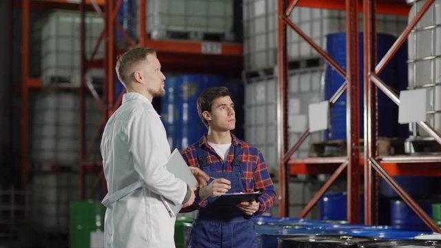 Tracking medium shot of middle aged enginner in white coat pointing to steel barrels placed on racks and giving directions to young warehouse worker taking notes in factory storehouse