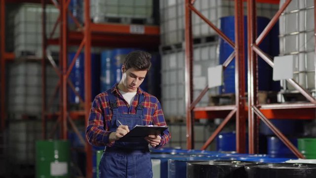 Tracking medium shot of young warehouse worker revising steel barrels on racks in factory storage and making notes in document on clipboard